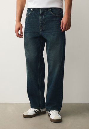 Person trägt dunkelblaue Jeans mit geradem Bein und weiße Sneaker mit schwarzen Streifen und steht auf grauem Boden.