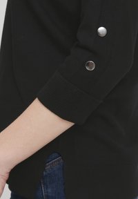 Juodas megztinis su minkštu audiniu ir raukšlėtais rankogaliais, su sidabriniais sagčių mygtukais ant rankovių ir šonuose įpjovimu, apsivilktas ant mėlynų džinsų.