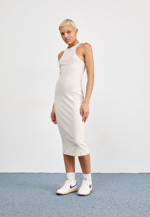 Robe côtelée midi, de couleur beige clair, avec un col haut et sans manches, associée à des baskets blanches et bleues.