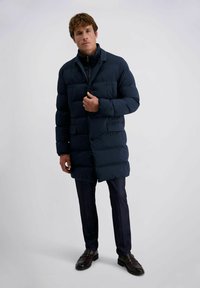 Homme portant un long manteau matelassé bleu marine, un pantalon bleu marine et des chaussures de ville noires, se tenant devant un fond clair et uni.