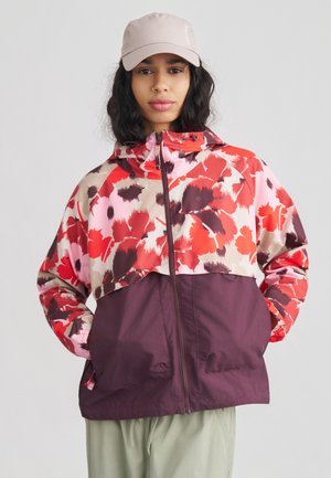 Ženska, oblečena v rdečo in rožnato jakno z vzorcem cvetja, spodnji del vijolične barve, in svetlo rožnatim klobukom, stoji z rokami v žepih.