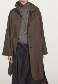 Brązowy, oversize'owy płaszcz z wysokim kołnierzem, z guzikiem i elastycznymi mankietami, noszony na szarym golfie i ciemnych spódnicach plisowanych.