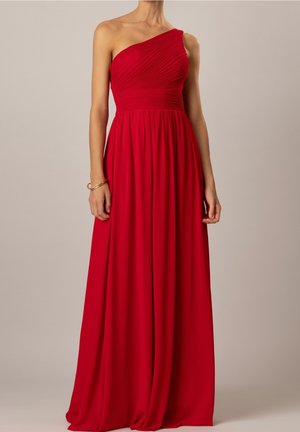 Femme portant une robe rouge longue jusqu'au sol, à une épaule, avec un corsage ajusté plissé et une jupe fluide, debout devant un fond uni.