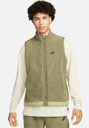 Green fleece vest with a high collar and zip closure, featuring a black Nike logo and a contrasting light green hem. Worn over a white long-sleeve shirt.