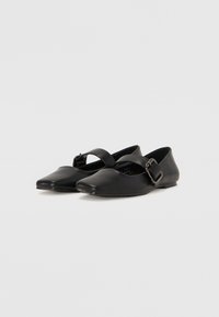 Des ballerines noires en cuir avec un bout pointu, présentant une sangle croisées et un détail de boucle métallique sur le côté. Texture lisse.