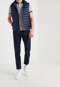 Marinblå puffer väst, grå kortärmad polo med krage, marinblå byxor och vita sneakers med texturerade sulor. Klara linjer och subtila varumärkesdetaljer synliga.