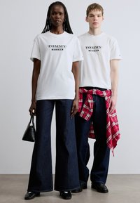 Λευκές μπλούζες με το λογότυπο "TOMMY JEANS", συνδυασμένες με σκούρα τζιν παντελόνια με καμπάνα. Ένα μοντέλο κρατάει μια μαύρη τσάντα, ενώ το άλλο φοράει μια κόκκινη καρό πουκάμισο δεμένη στη μέση.