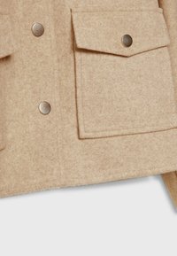 Veste en laine beige en gros plan montrant un tissu texturé, deux boutons-pression métalliques et une grande poche avant à rabat avec fermeture pression.