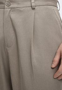 Κοντινό πλάνο σε μπεζ παντελόνι με πτυχώσεις μπροστά, κούμπωμα με κουμπί, πλαϊνή τσέπη και χέρι που ακουμπά κοντά στην δεξιά τσέπη.