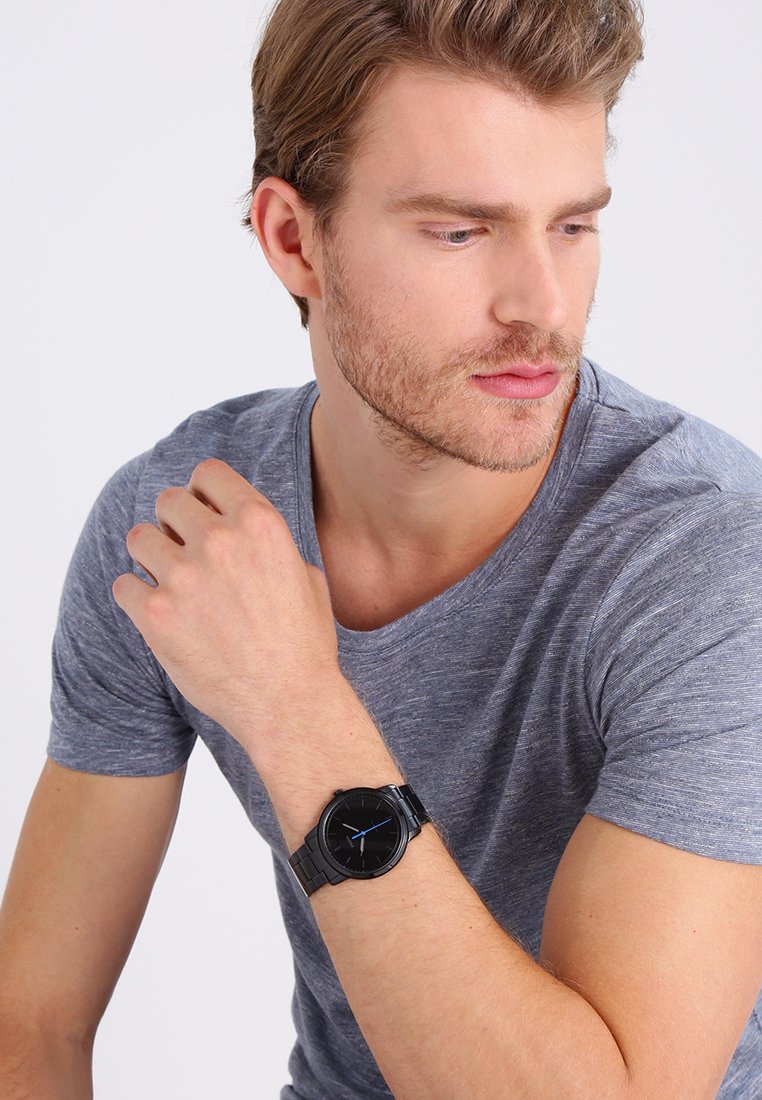 Reloj analógico negro con acentos azules, cara redonda y correa de metal negro texturizado; lleva puesto en la muñeca izquierda de una persona que viste una camiseta gris.
