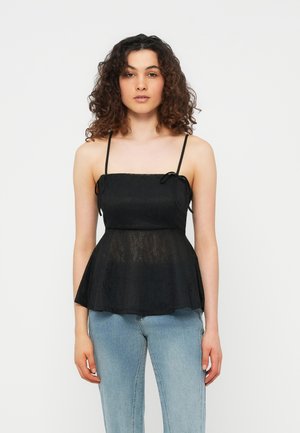 Vrouw met krullend haar, gekleed in een zwart mouwloos topje met dunne bandjes en lichtblauwe spijkerbroek, staand voor een effen witte achtergrond.