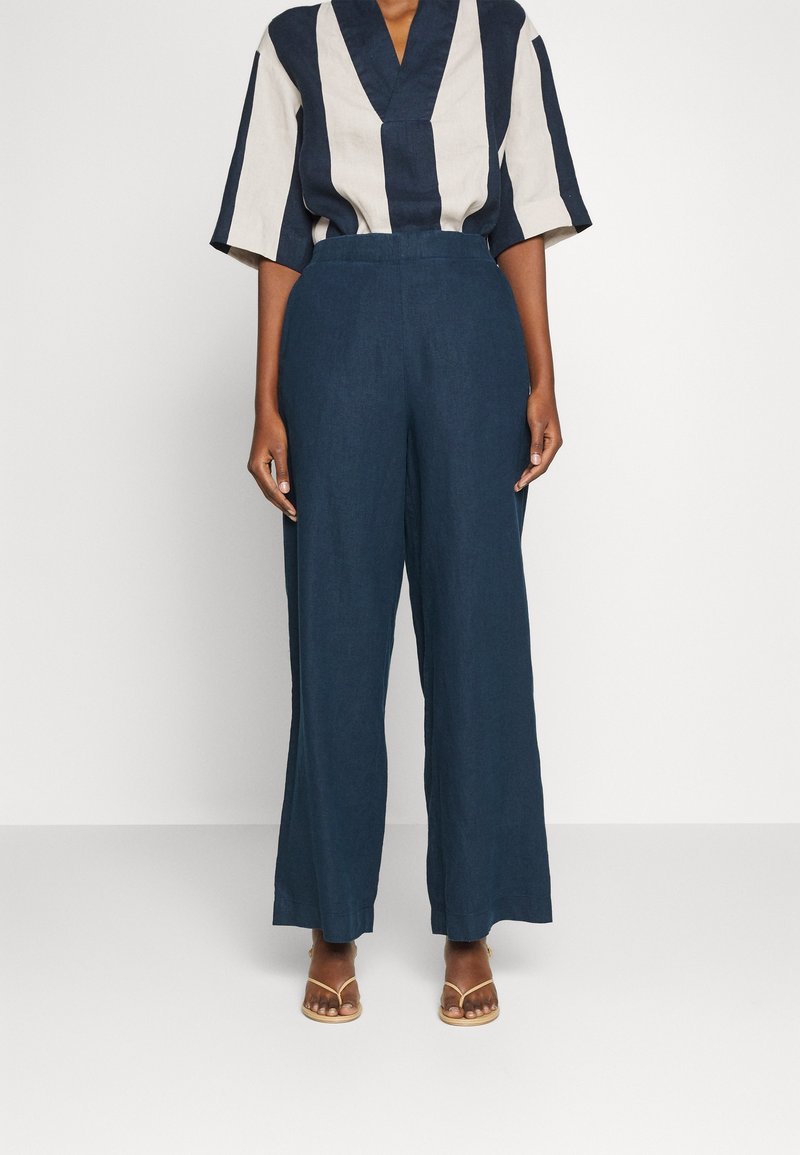Personne portant un pantalon large marine, un haut à col en V rayé beige et marine, et des sandales beige à bouts ouverts, debout devant un fond blanc.