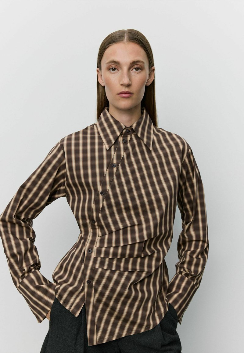Femme aux longs cheveux raides portant une chemise à carreaux marron à boutons avec des plis asymétriques et un pantalon noir sur un fond clair uni.
