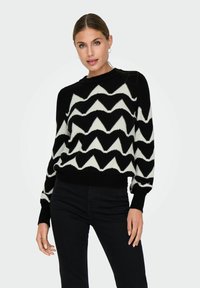 Maglione a righe zigzag bianco e nero, con scollo rotondo e polsini e orlo a costine, indossato con pantaloni neri. Texture morbida.