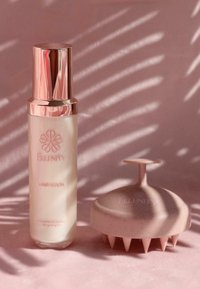 Siero per capelli in una bottiglia trasparente con un cappuccio dorato, accanto a un massaggiatore per cuoio capelluto rosa con bordi appuntiti, entrambi su una superficie morbida rosa.