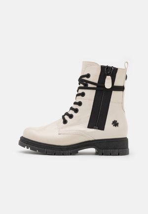 Beżowy skórzany wojskowy but z czarnymi sznurówkami, bocznym zamkiem błyskawicznym, czarną podeszwą i małym detalem koniczyny czterolistnej na obcasie.