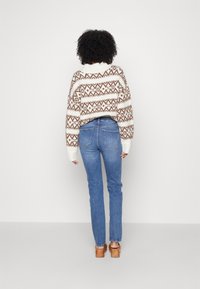 Suéter de crochet con un patrón geométrico marrón, base blanca y ajuste oversize; combinado con jeans azules de pierna recta y zapatos de tacón color beige.