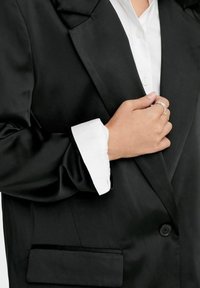 Blazer en satin noir avec revers crantés, fermeture à un bouton et une poche poitrine. Poignets de chemise blancs visibles, affichant une finition nette.