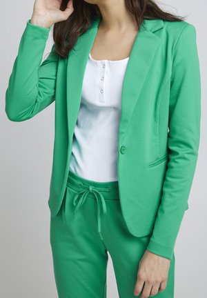 Femme portant un blazer vert et un pantalon assorti avec cordon de serrage, accompagné d'un chemisier blanc côtelé à boutons, sur fond uni.