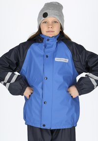 Blå og marineblå vandtæt jakke med elastiske manchetter, knaplukning og reflekterende striber. Sammen med en grå strikket beanie.