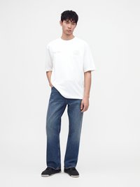 Νέος άνδρας που φοράει λευκό oversized t-shirt, μπλε χαλαρά τζιν και μαύρα αθλητικά παπούτσια, στέκεται μπροστά σε απλό λευκό φόντο.