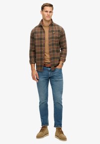 Camicia flanella a quadri nei toni del verde, marrone e rosso, indossata sopra un maglione beige, abbinata a jeans blu e stivali beige con lacci. Cintura marrone.