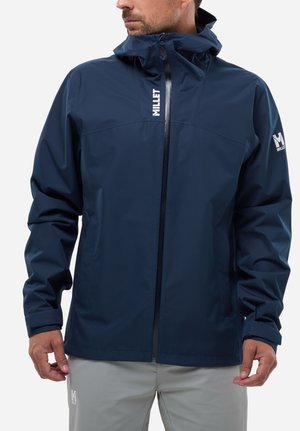 Man wearing navy blue waterproof jacket with hood and zipper, showing Millet brand logos on chest and sleeve, paired with light gray pants.