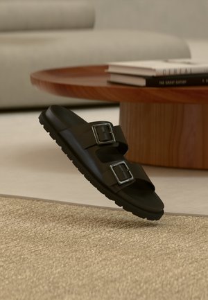 Sandale noire à talon avec deux boucles argentées ajustables, reposant sur un tapis beige texturé, près d'une table basse ronde en bois avec des livres empilés.