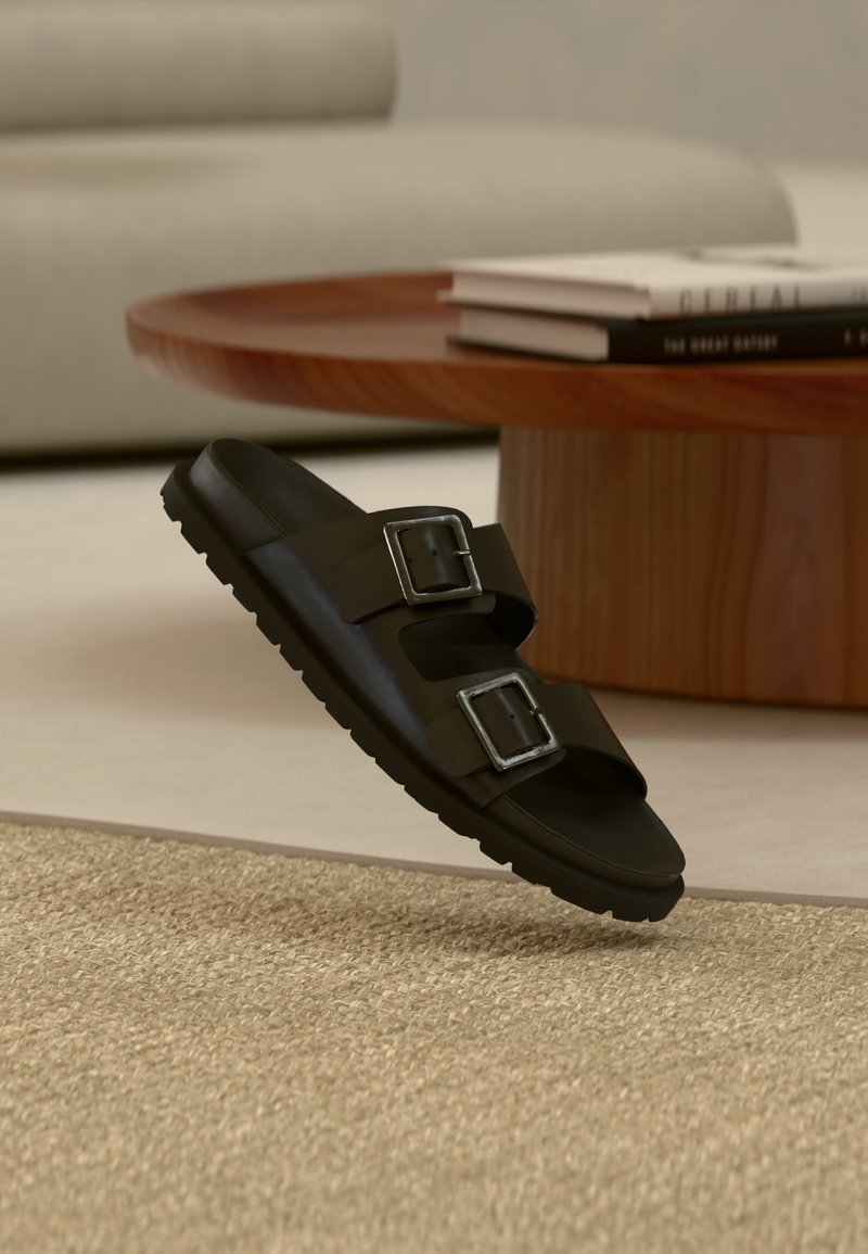 Sandale noire à talon avec deux boucles argentées ajustables, reposant sur un tapis beige texturé, près d'une table basse ronde en bois avec des livres empilés.