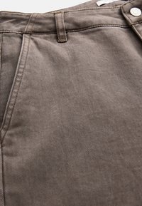 Nærbillede af brun denimmateriale, der viser lomme, bæltestrop og sølvsnap på linningen af bukser.