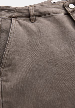 Lähikuva ruskeasta denim-kankaasta, jossa näkyy tasku, vyölenkki ja hopeinen nappi housujen vyötäröllä.