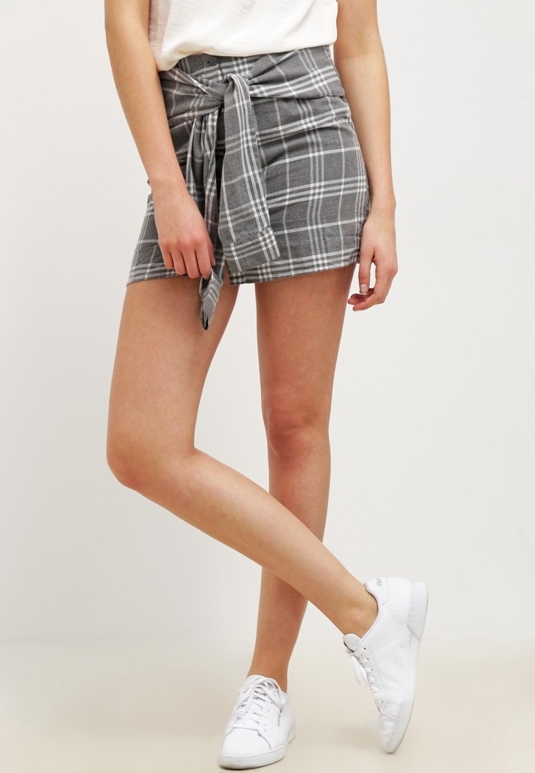Femme avec les jambes croisées portant une mini-jupe grise à carreaux avec un détail de nœud devant, un top blanc et des baskets blanches, debout devant un fond uni.