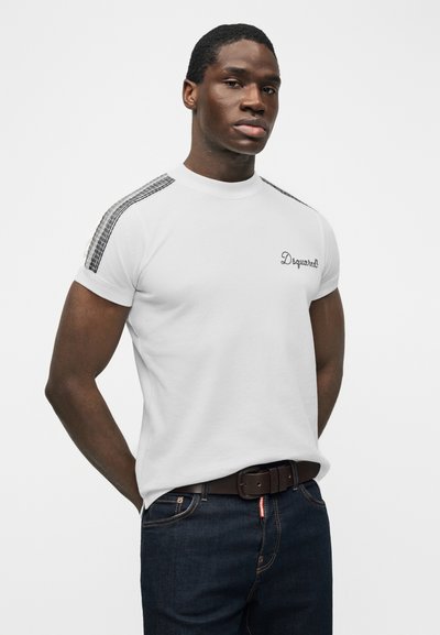 Μαύρος άνδρας στέκεται με τα χέρια πίσω από την πλάτη, φορώντας λευκό κοντομάνικο πουκάμισο με μαύρες ρίγες στους ώμους και σκούρα τζιν με καφέ ζώνη.