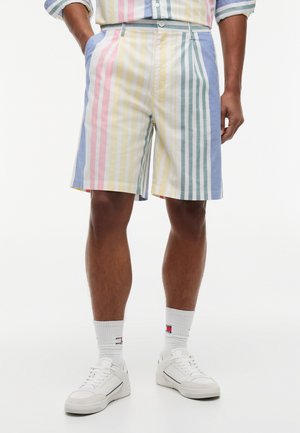 Mehrfarbige, vertikal gestreifte Shorts, getragen von einer Person, die mit den Händen in den Taschen steht, kombiniert mit weißen Sneakern und markenbestickten Crew-Socken.