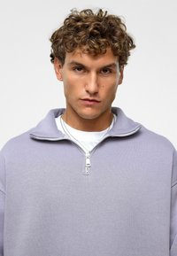Lichtpaarse fleece sweatshirt met rits en hoge kraag, gemaakt van zachte stof. Gelaagd over een wit shirt, model kijkt naar voren.