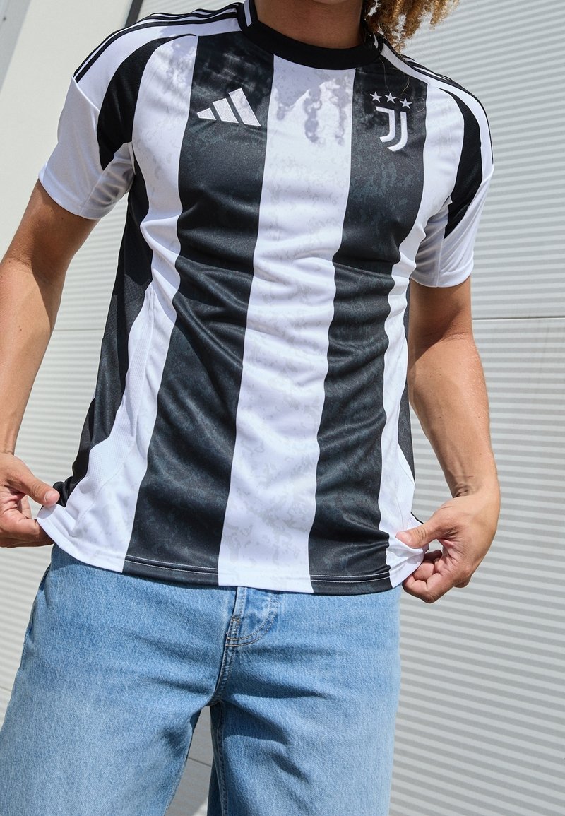 Personne portant un maillot de football rayé noir et blanc de la Juventus et un jean bleu, tenant le maillot par les côtés.