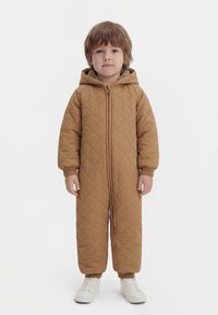 Jeune enfant debout, portant une combinaison matelassée à capuche marron avec fermeture éclair et des baskets blanches, sur un fond blanc uni.