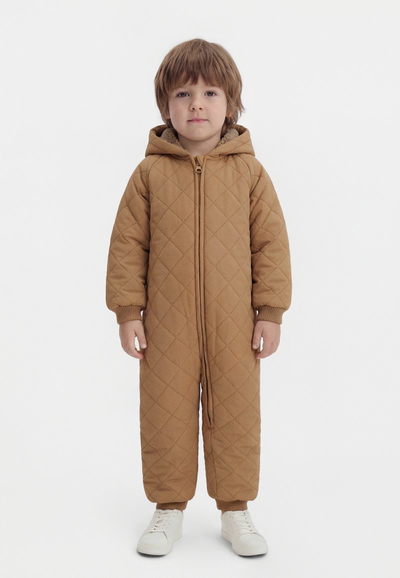 Jeune enfant debout, portant une combinaison matelassée à capuche marron avec fermeture éclair et des baskets blanches, sur un fond blanc uni.