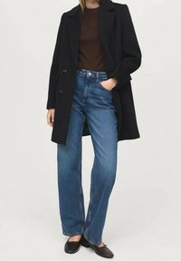 Femme portant un manteau noir, un haut marron, un jean bleu taille haute à jambes larges et des ballerines noires, debout avec une main dans la poche.