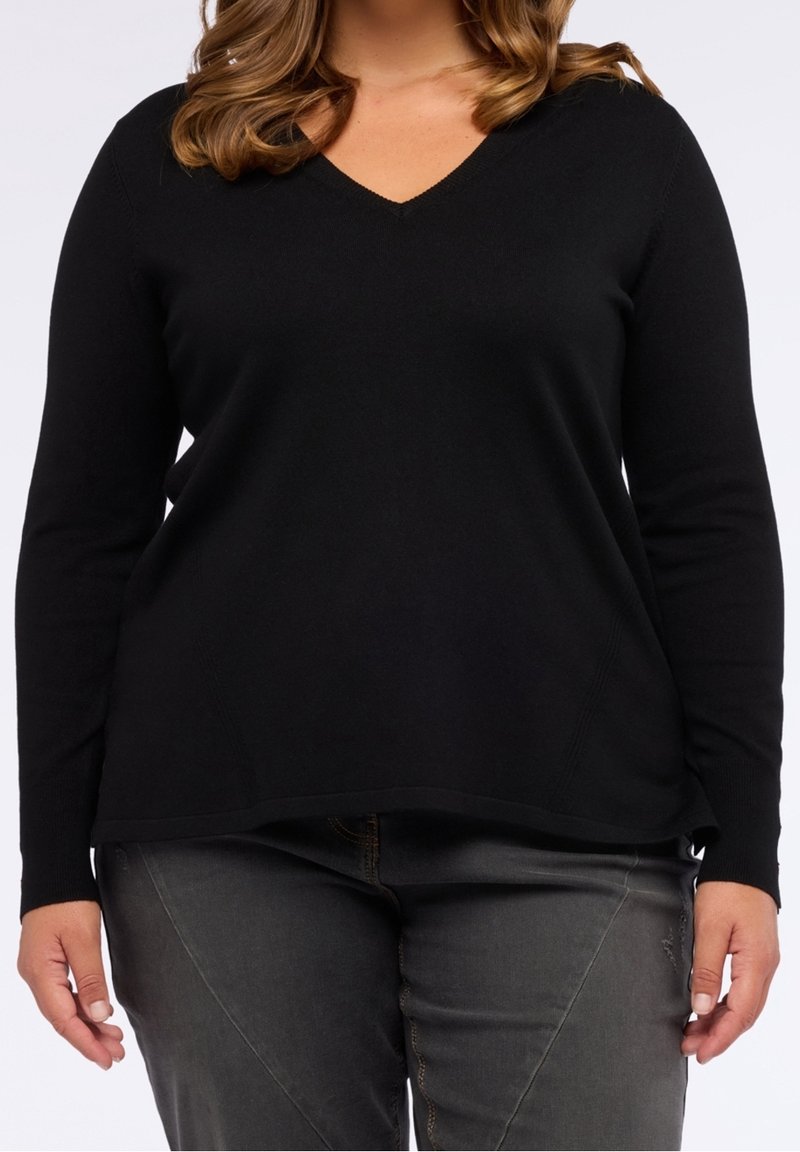 Femme portant un pull noir à col en V manches longues et un jean gris foncé, debout avec les bras détendus le long du corps devant un fond uni.
