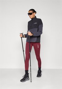 Giacca zip-up nera e grigia con colletto alto, leggings amaranto e scarpe da trekking nere. Tiene in mano dei bastoni da trekking e indossa occhiali da sole.