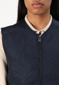 Marineblauwe gewatteerde vest met geometrisch reliëfpatroon, volledige rits aan de voorkant, soepele textuur en geribbelde beige kraag eronder.