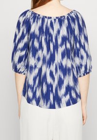 Blusa blu e bianca con stampa, maniche a palloncino, scollatura elasticizzata e vestibilità rilassata. Il tessuto sembra leggero con una texture liscia.
