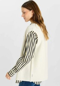 Maglione girocollo bianco in maglia sopra una camicia a righe in bianco e nero. Presenta una texture a coste e una silhouette dritta con uno spacco laterale.
