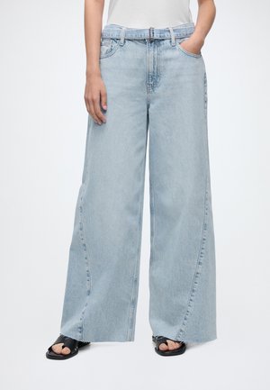 Jean bleu clair à jambes larges avec une ceinture en denim intégrée, porté avec des sandales noires à bout ouvert et un haut blanc.