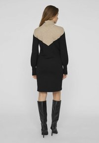 Robe en tricot avec un col montant ; corps noir et zone d'épaule beige, arborant un design en V. Manches longues et bouffantes, et silhouette ajustée.