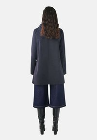 Lungo cappotto blu navy con un taglio dritto, abbinato a culotte in denim a gamba larga e stivali neri fino al ginocchio. La texture è liscia e contemporanea.