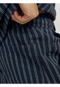 Tamsiai mėlynas tekstūruotas audinys su baltomis vertikaliomis juostelėmis; arti pavaizduota kišenė ir liemuo, rodanti laisvo pritaikymo dizainą.