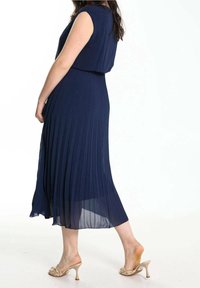 Robe plissée bleu marine avec un corsage cintré et une jupe fluide, confectionnée en tissu léger. Associée à des sandales à talons beiges.