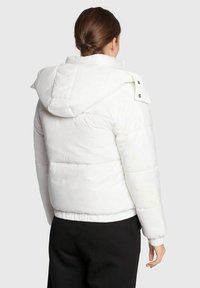 Chaqueta de plumas blanca con capucha, que presenta un diseño acolchado, puños elásticos y una cintura ajustada. Decorada con botones a presión en el cuello.